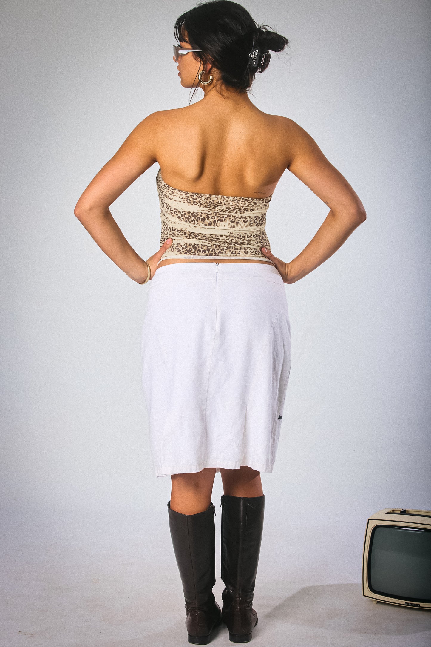 Woman wearing a strapless top and white skirt with black boots, standing against a plain background.