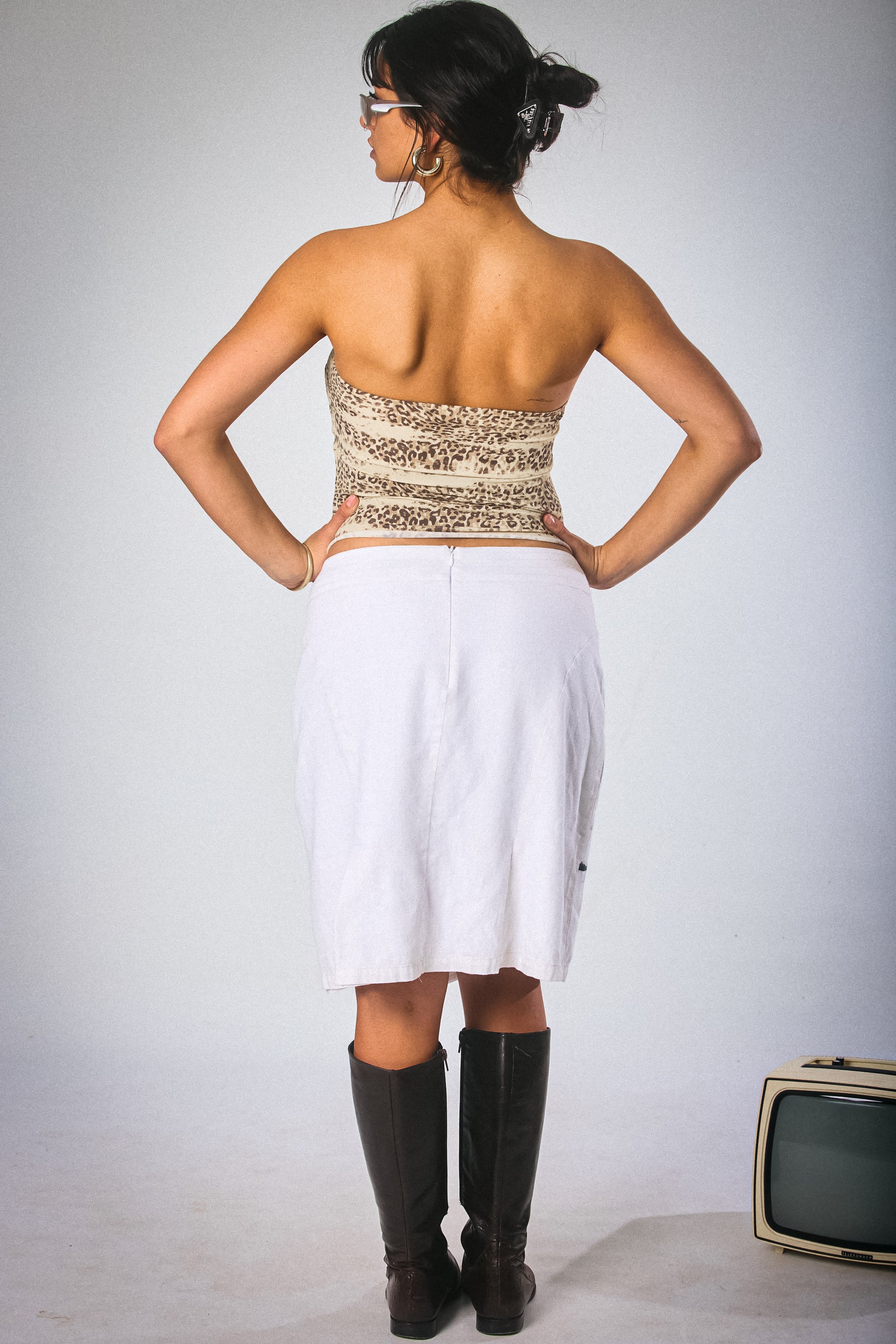 Woman wearing a strapless top and white skirt with black boots, standing against a plain background.