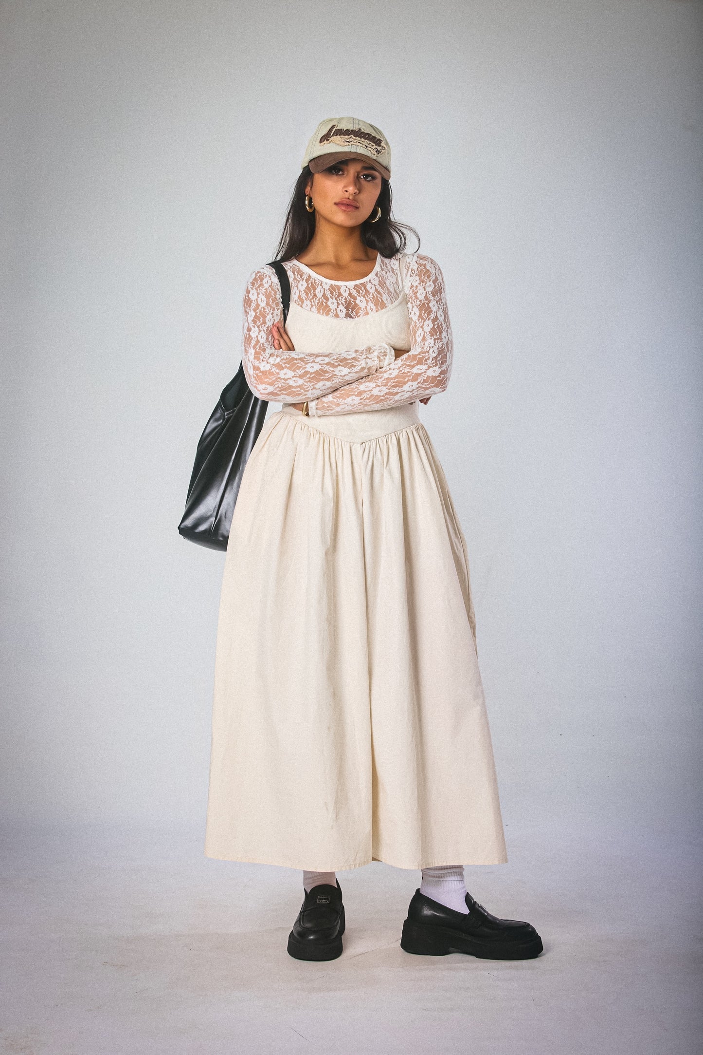 Person wearing a white lace top, beige skirt, black shoes, and a black bag on a plain background